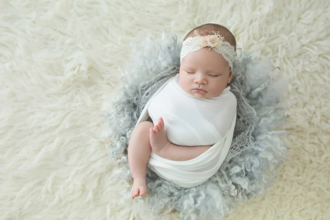 A sleeping baby girl wrapped in a white cloth, wearing a lace headband with a flower, resting on a soft, fluffy cream-colored blanket.
