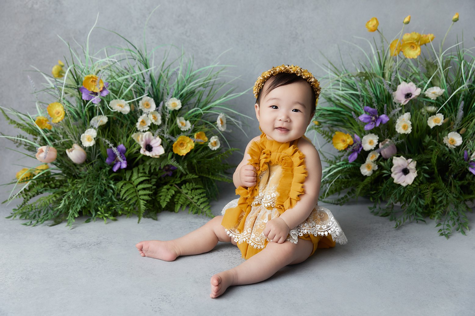 A smiling baby girl sitting on a gray surface with colorful flower arrangements in the background. She is wearing a mustard yellow dress with lace details and a matching headband.