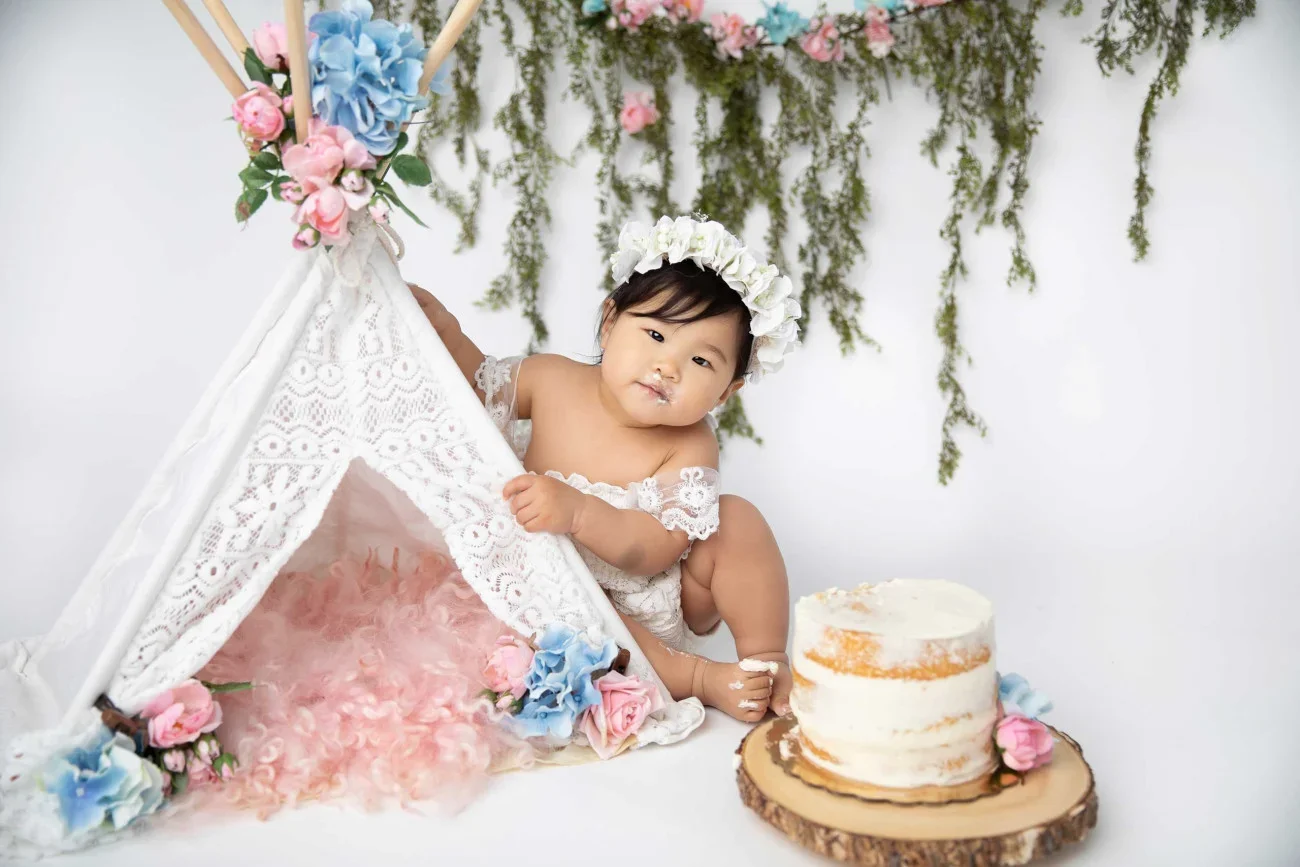 A young child with a flower crown is sitting beside a cake and a small tent decorated with flowers, with green foliage in the background.