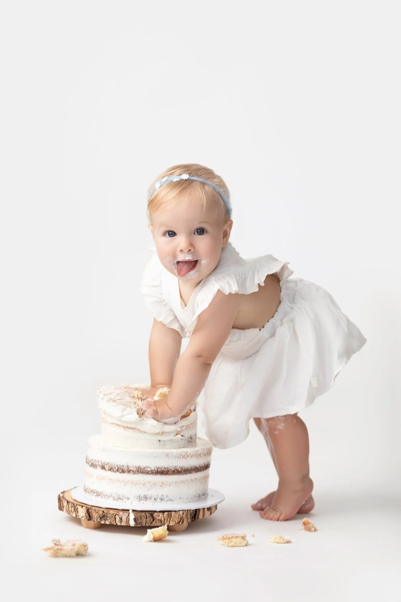 A young child with a blue headband and white dress playing with a small, layered cake on a wooden cake stand, with cake crumbs around her on a white background.