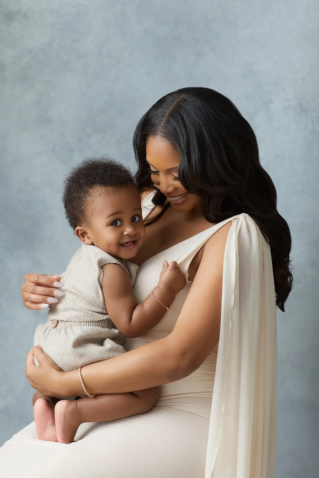 A woman holding a smiling toddler girl in her arms, with a gray background.