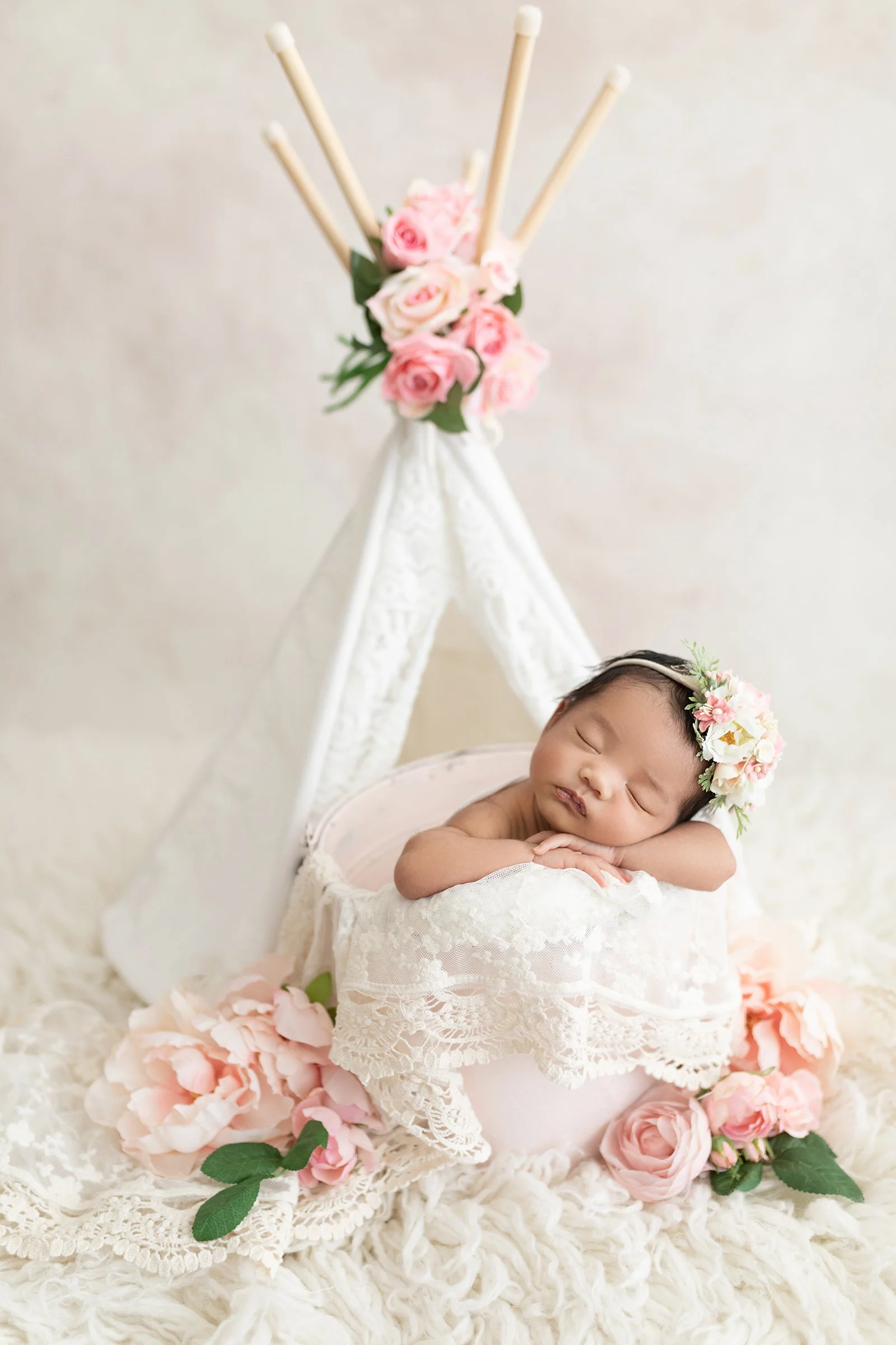 A sleeping baby with a floral headband resting in a lace-lined basket, surrounded by pink roses and peonies, with a decorative white lace teepee in the background.