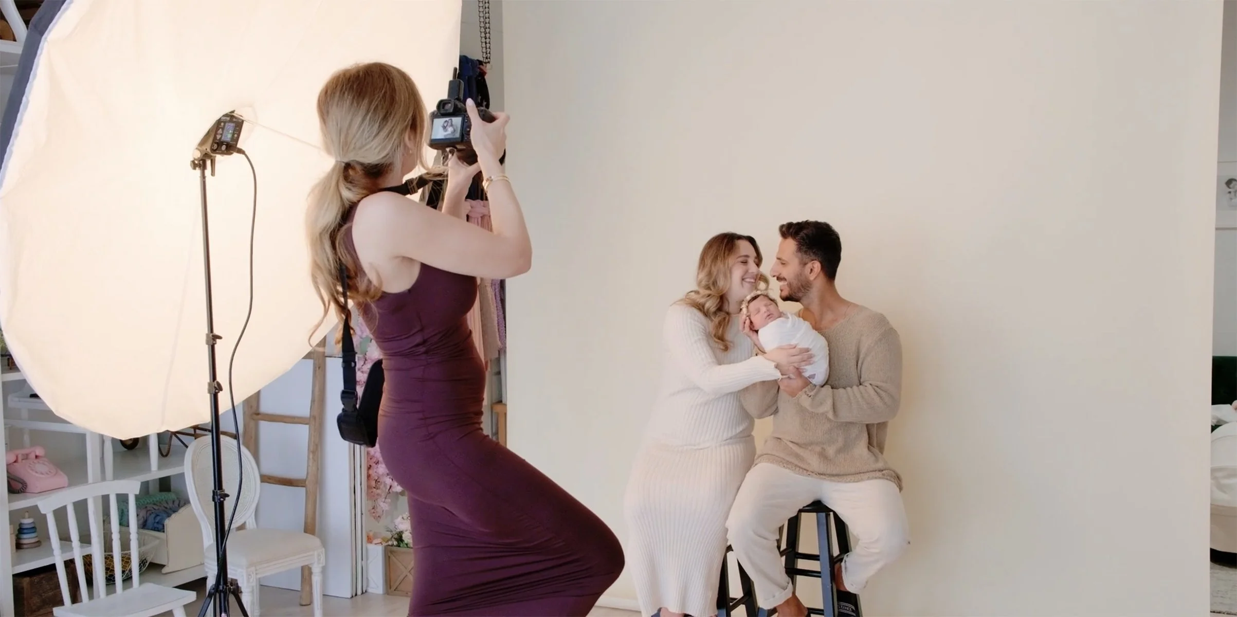 A mother and father holding their newborn baby during a photoshoot, with a photographer taking pictures against a plain background.