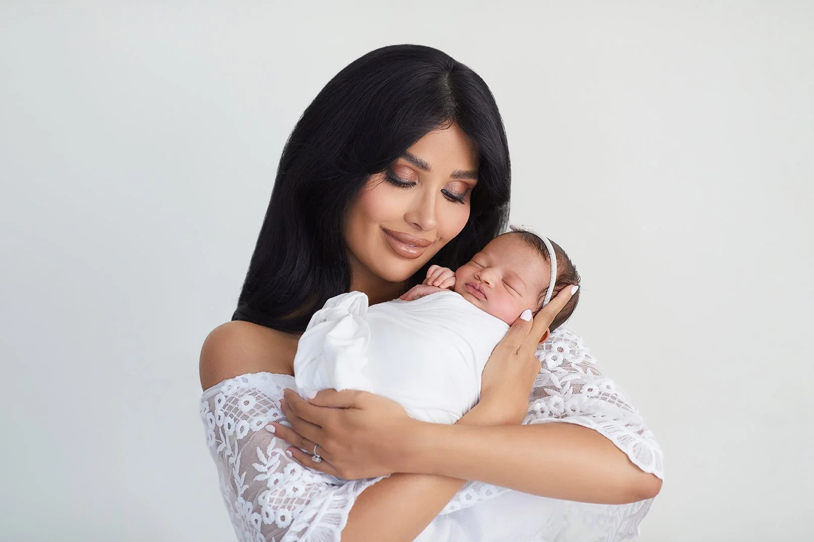 A woman with long dark hair and makeup holding a newborn baby with a headband, dressed in white, against a plain light background, smiling with closed eyes.