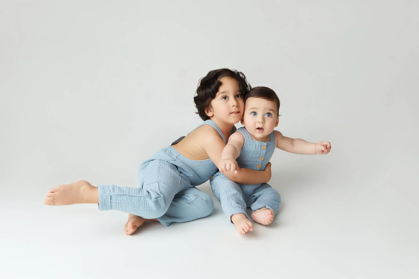 Two young children, a girl and a baby boy, sitting on the floor against a plain background, wearing matching light blue outfits. The girl has curly dark hair and is holding the boy. The boy has short dark hair and is reaching out with his arm.