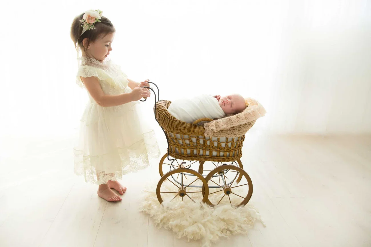 A young girl in a white dress with a floral headband pushes a vintage wicker baby stroller with a sleeping infant wrapped in white cloth.