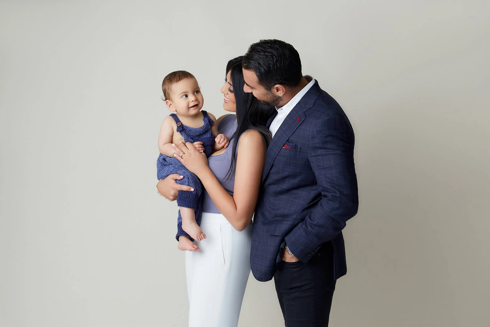 A family of three with a woman holding a young child and a man standing close by, smiling at the child, against a plain light-colored background.