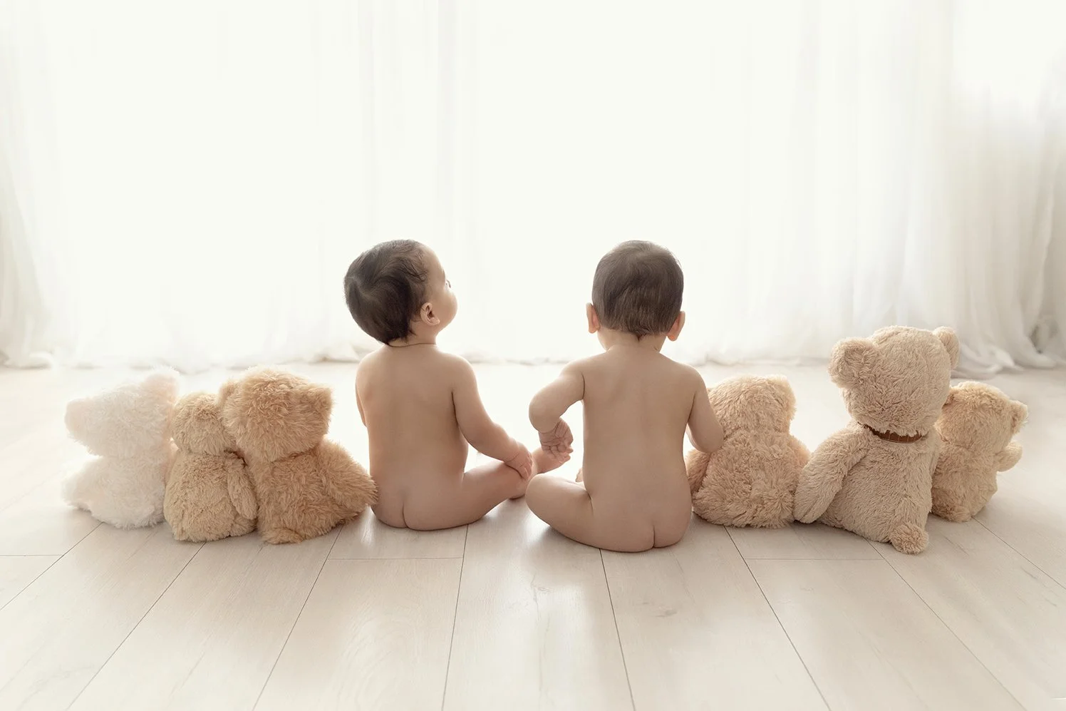 Two naked babies sitting on the floor with teddy bears, holding hands, facing a bright window with white curtains.