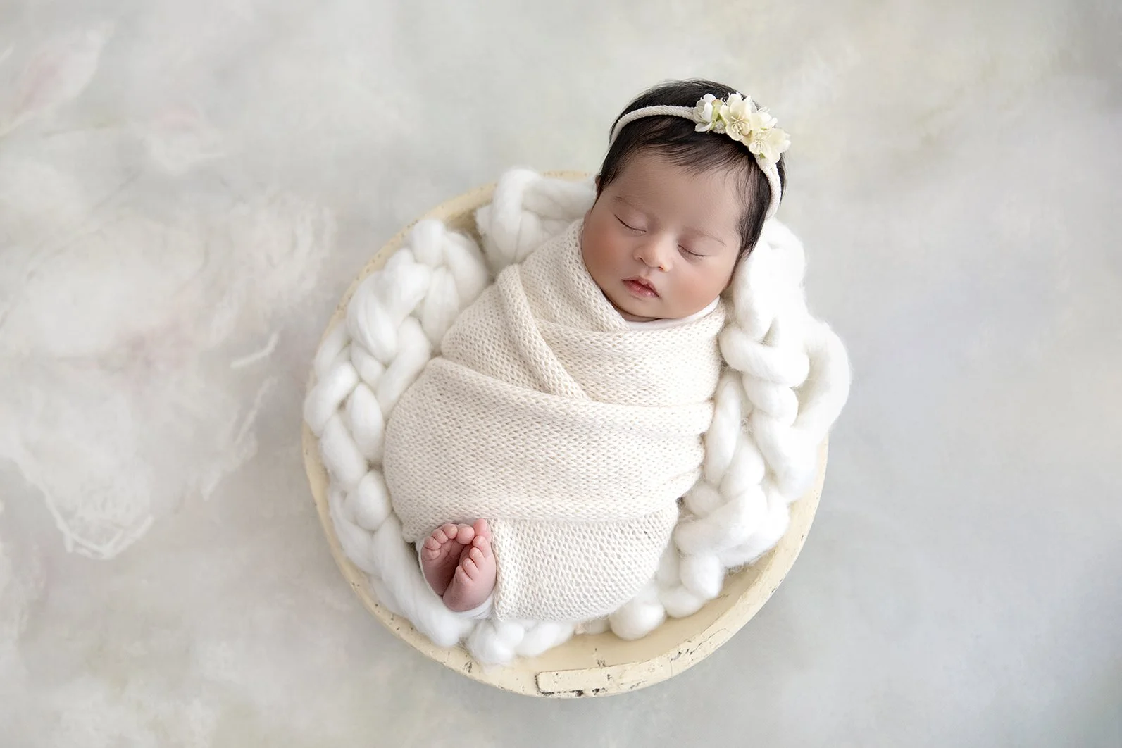 A sleeping baby girl swaddled in a cream blanket, lying on a fluffy white blanket in a round bowl, wearing a floral headband.