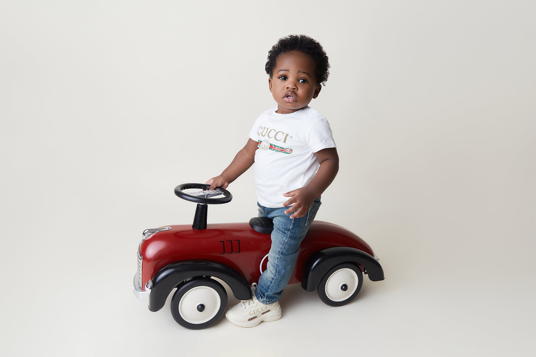 Young child with curly hair in a white Gucci t-shirt, blue jeans, and white sneakers, sitting on a red and black toy car against a plain white background.
