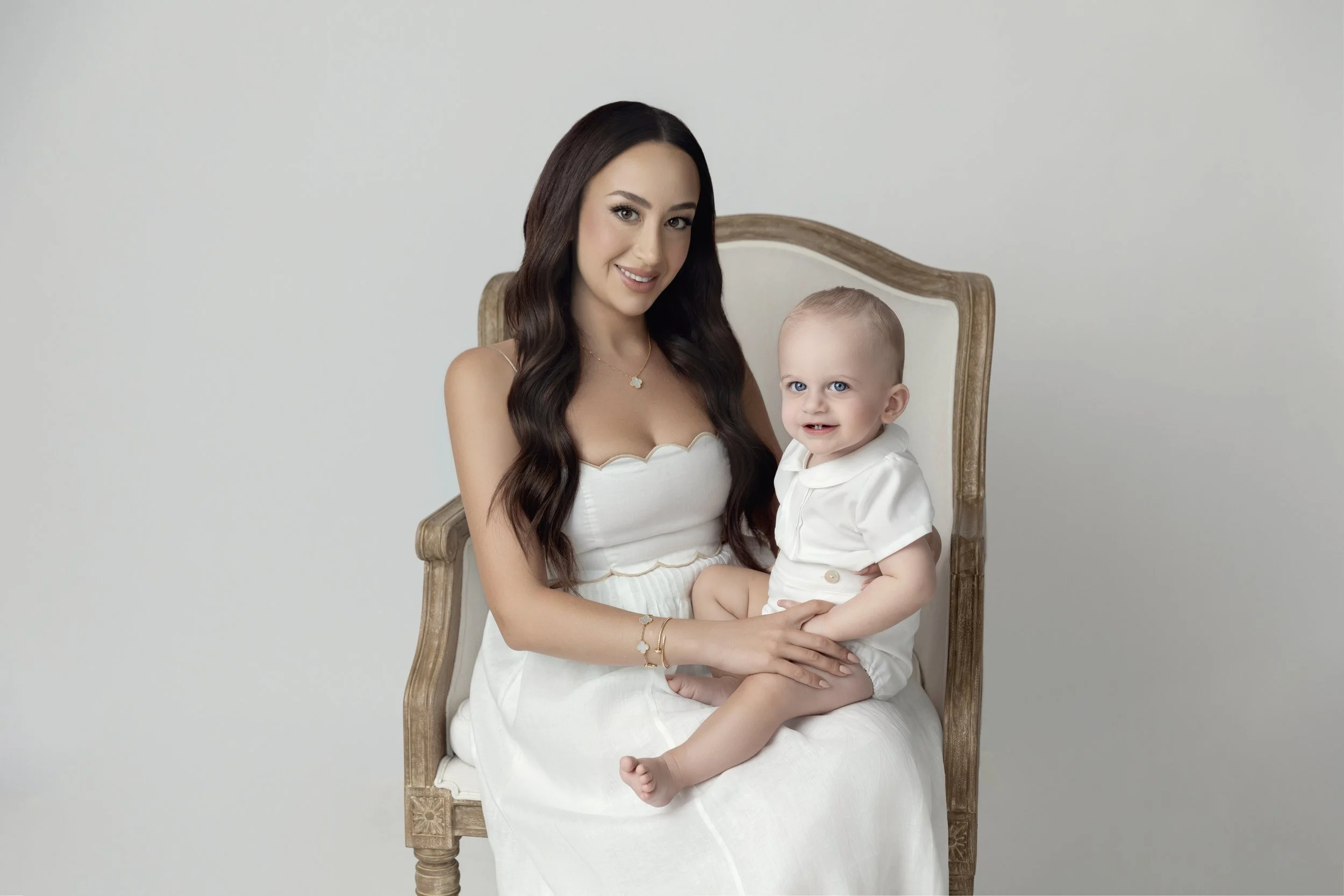 A woman with long dark hair smiling, holding a young boy with blond hair and blue eyes, sitting on a vintage wooden chair against a plain light background.