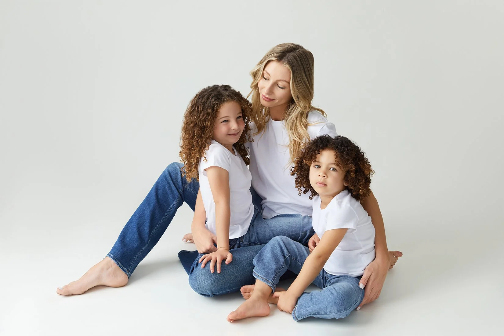 A woman with long, wavy blonde hair and two young girls with curly brown hair sitting together on a plain white background. They are dressed in white shirts and blue jeans, appearing happy and relaxed.