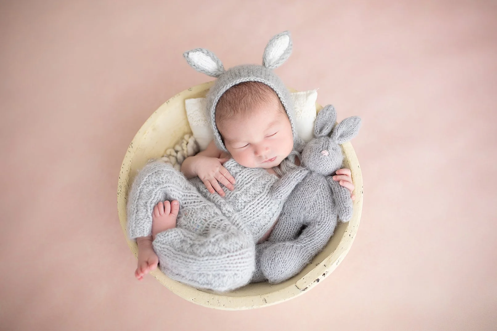 A sleeping baby in a cream-colored bowl wearing a gray knitted hat with bunny ears, holding a gray stuffed bunny, surrounded by soft blankets, on a light pink background.