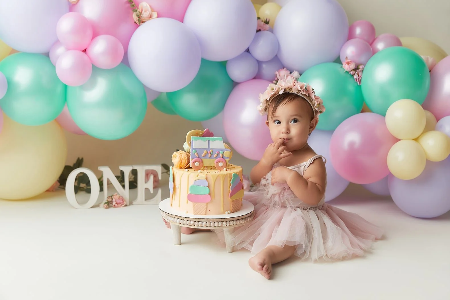 A young girl in a pink tutu dress with a floral headband sits next to a cake and colorful balloons, celebrating a first birthday.