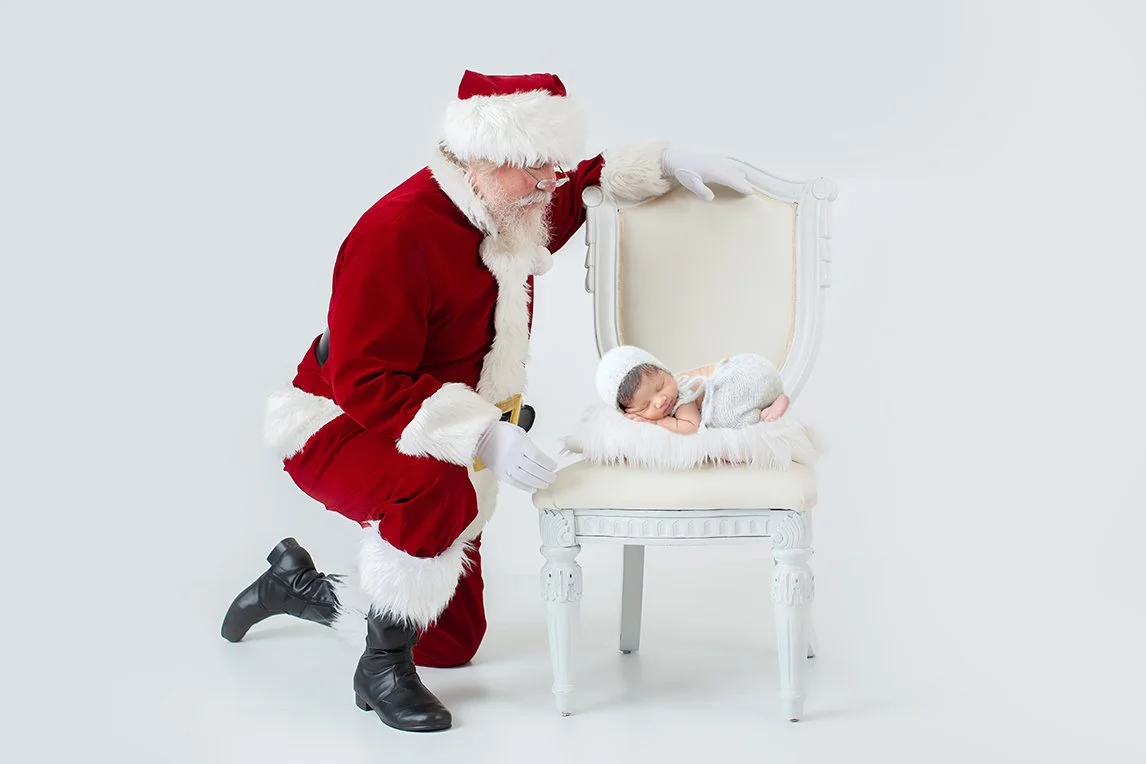 Santa Claus kneeling beside a white chair with a newborn baby sleeping on it, set against a plain white background.