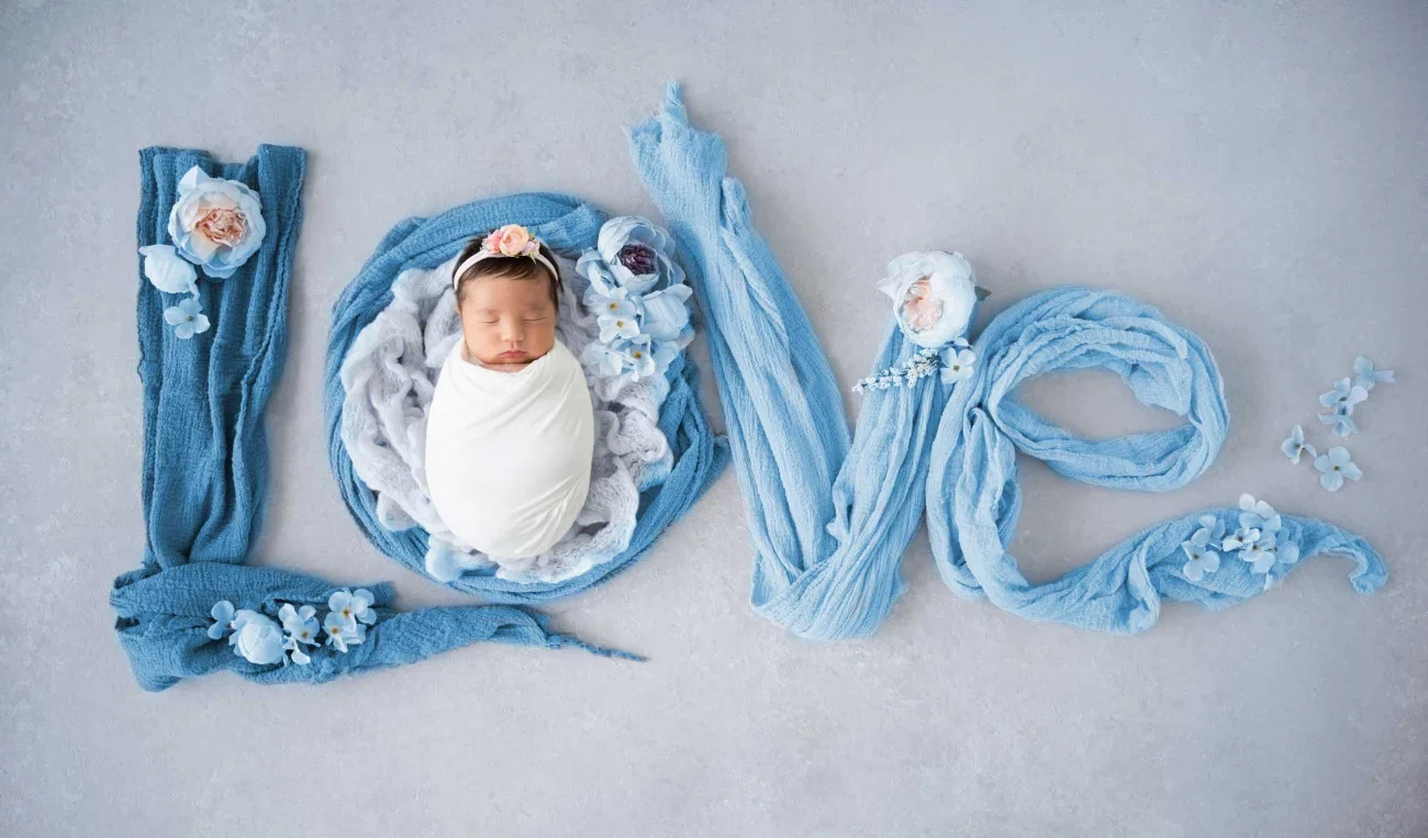 Swaddled baby sleeping with the word "love" spelled out in blue fabric and flowers.