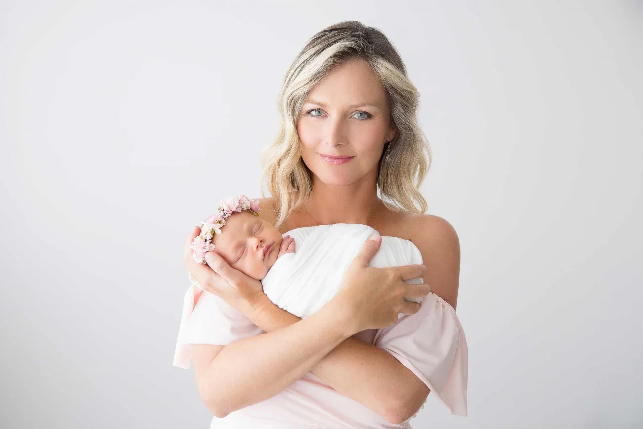 A woman with blonde hair holding a newborn baby, wrapped in a white blanket and wearing a pink floral headband, against a plain white background.