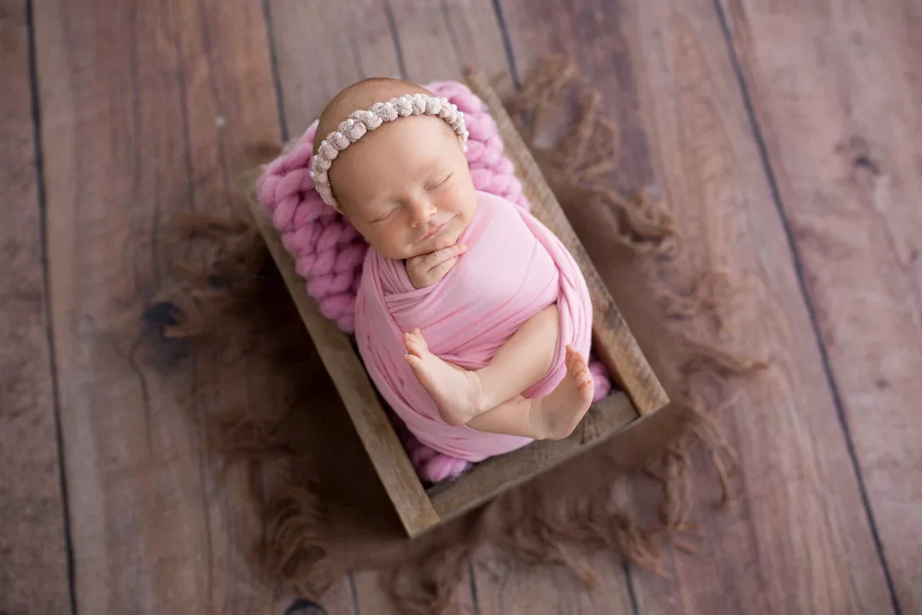 A sleeping baby girl wrapped in pink swaddle, wearing a pink headband with beads, resting on a pink blanket inside a small wooden crate on a wooden floor.