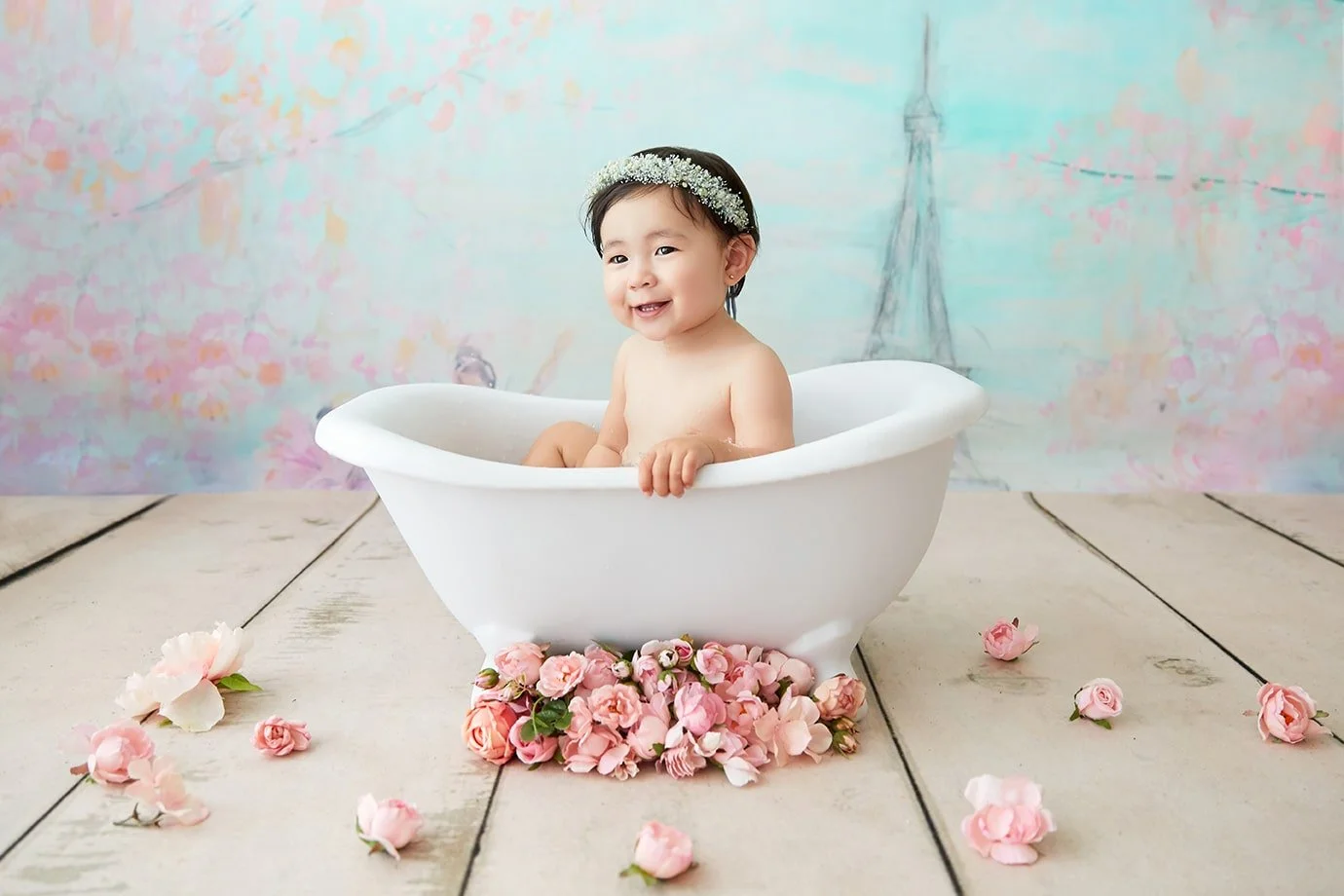A smiling baby girl with a floral headband sitting in a white bathtub surrounded by scattered pink flower petals and a pile of pink roses underneath, with a pastel-colored backdrop featuring faint drawings of flowers and the Eiffel Tower.