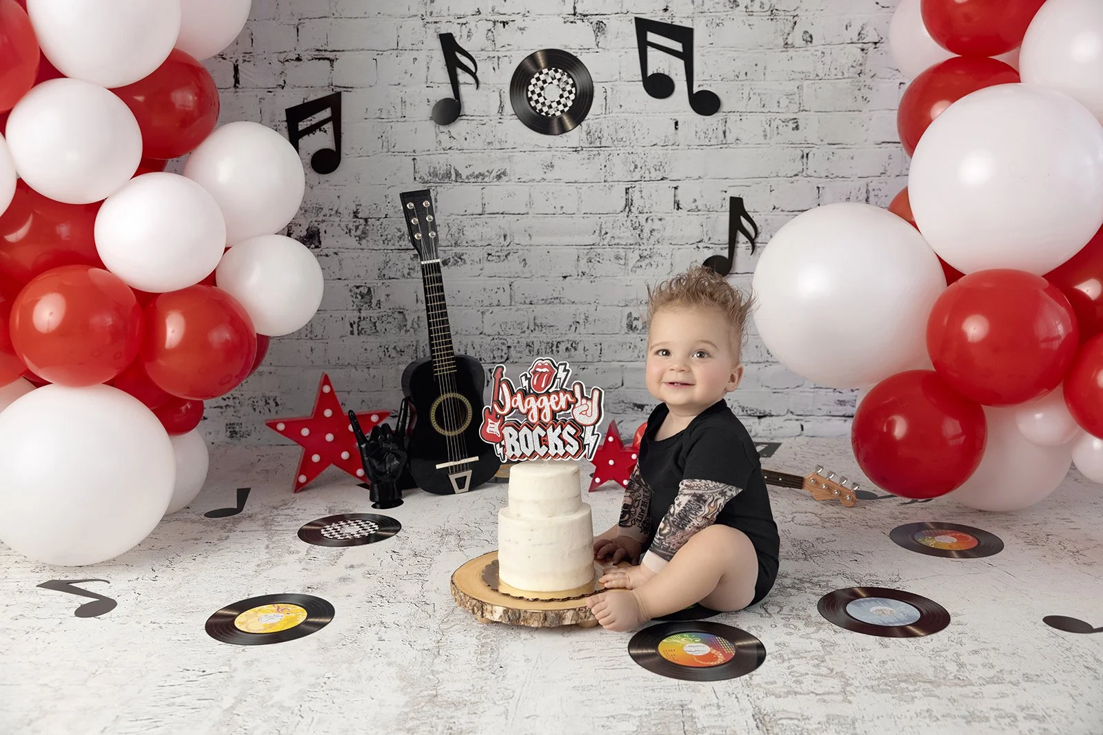 A birthday celebration with a young child sitting on the floor next to a white cake, decorated with musical-themed decorations including vinyl records, guitars, and note cutouts, with balloons and a brick wall backdrop.