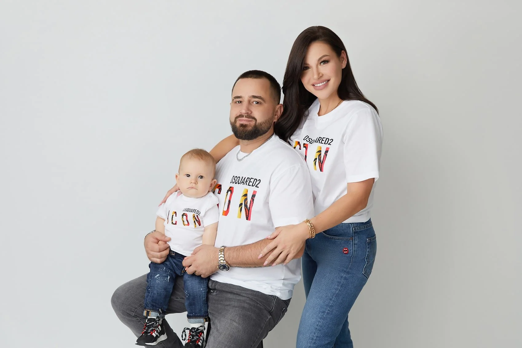 Family portrait of three: a man holding a young child, and a woman standing beside them. All are wearing white t-shirts with colorful text.