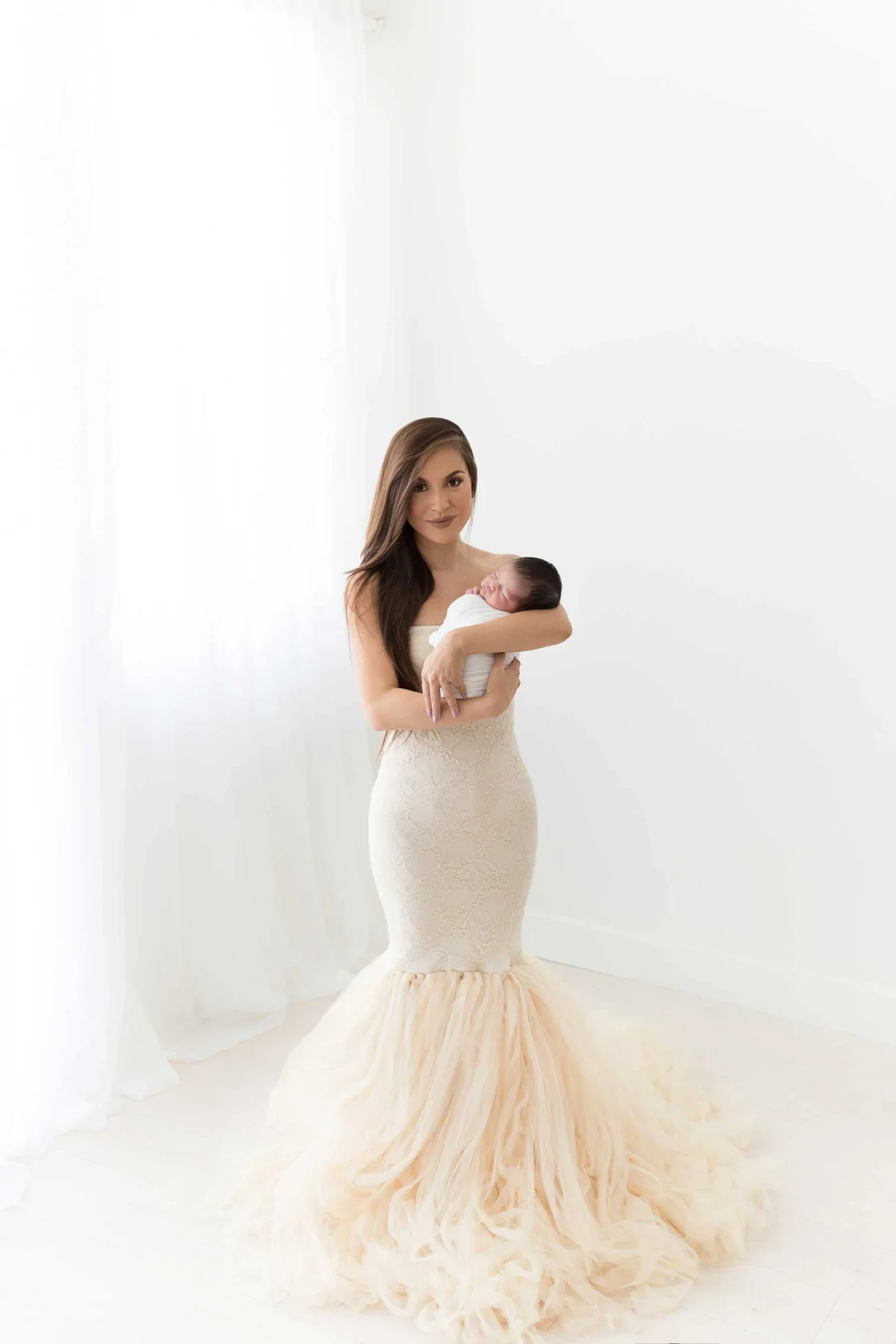 A woman in a white wedding dress holding a newborn baby in a minimalistic white room.