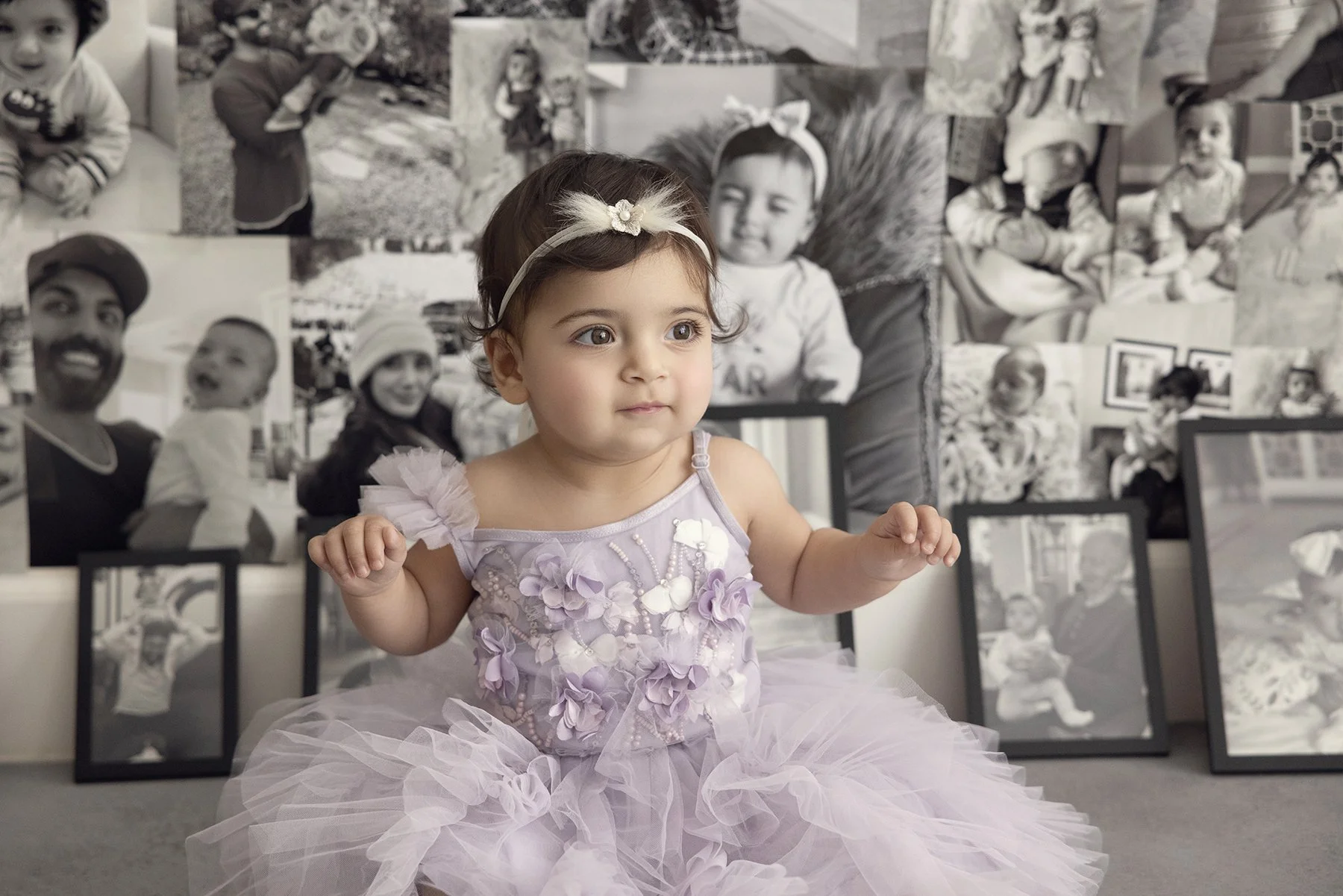 A young girl wearing a lavender dress with floral embellishments and a matching headband is sitting on the floor in front of a collage of black-and-white photographs of various people.