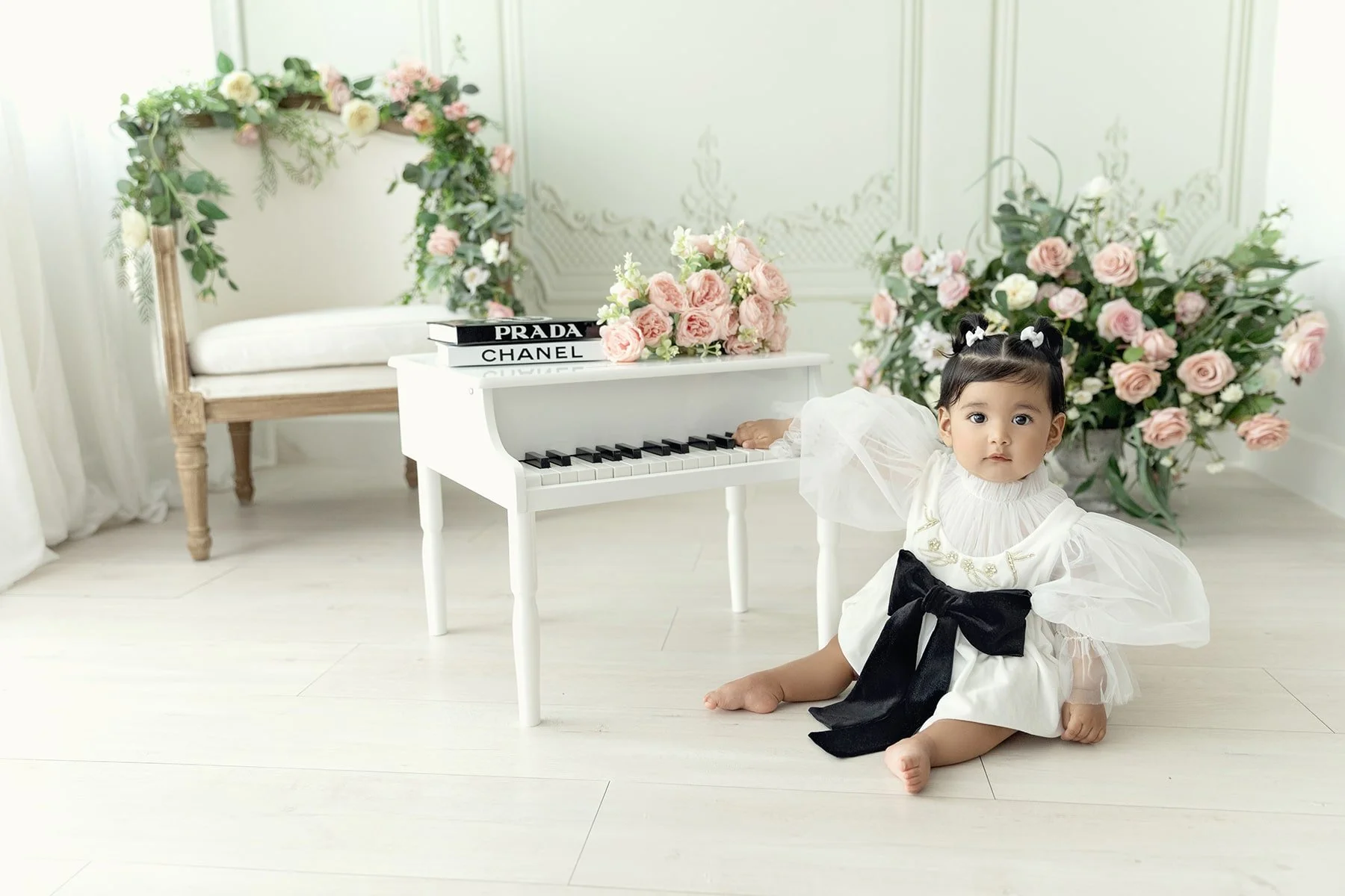 A young girl wearing a white dress with puffy sheer sleeves, a large black bow, and decorative gold embroidery, sitting on a white wooden floor in a room decorated with pink and white roses, a white toy grand piano, and a floral arch in the backgroun