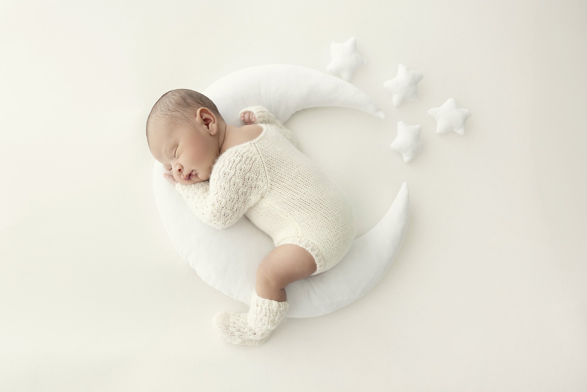 A sleeping baby wearing a knitted romper and booties, lying on a crescent moon-shaped pillow surrounded by small star-shaped pillows on a white background.