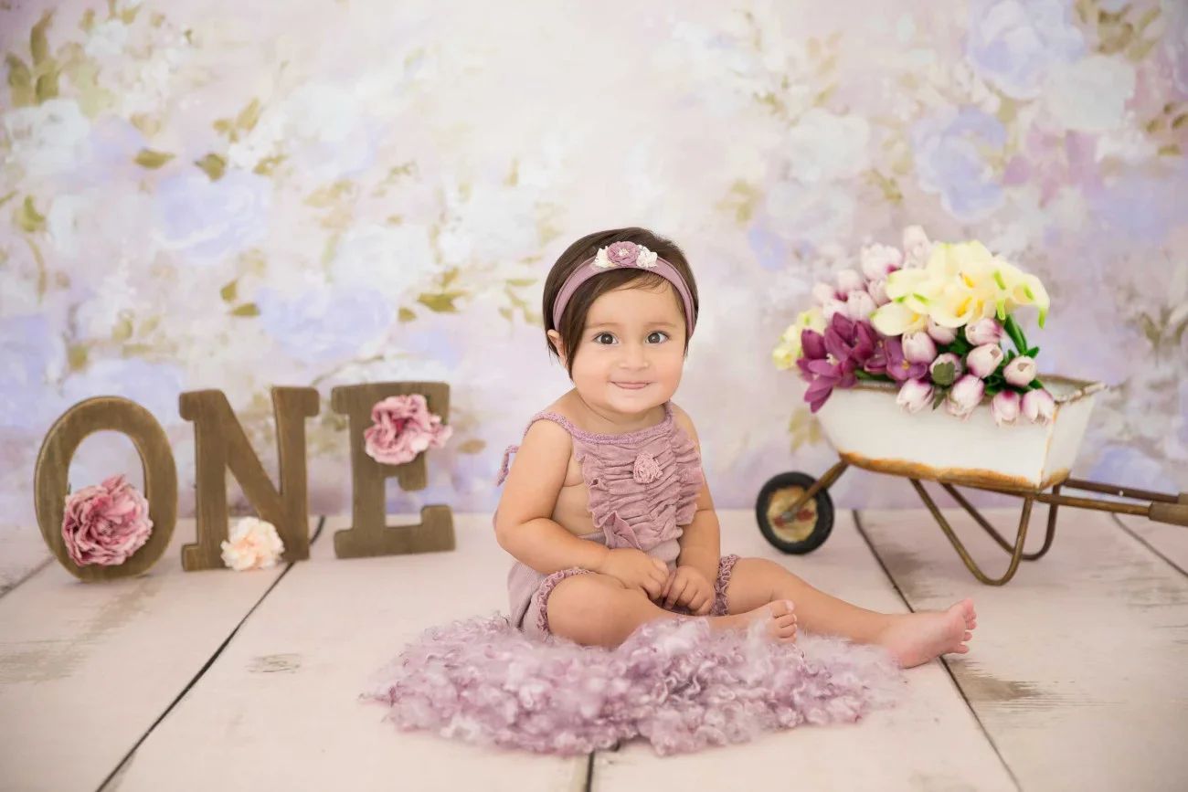 A baby girl sitting on a light-colored wooden floor in front of a pastel floral backdrop. She is wearing a pink ruffled romper and a matching headband. Behind her, large decorative letters spell 'ONE' with pink flowers, and there is a small white whe