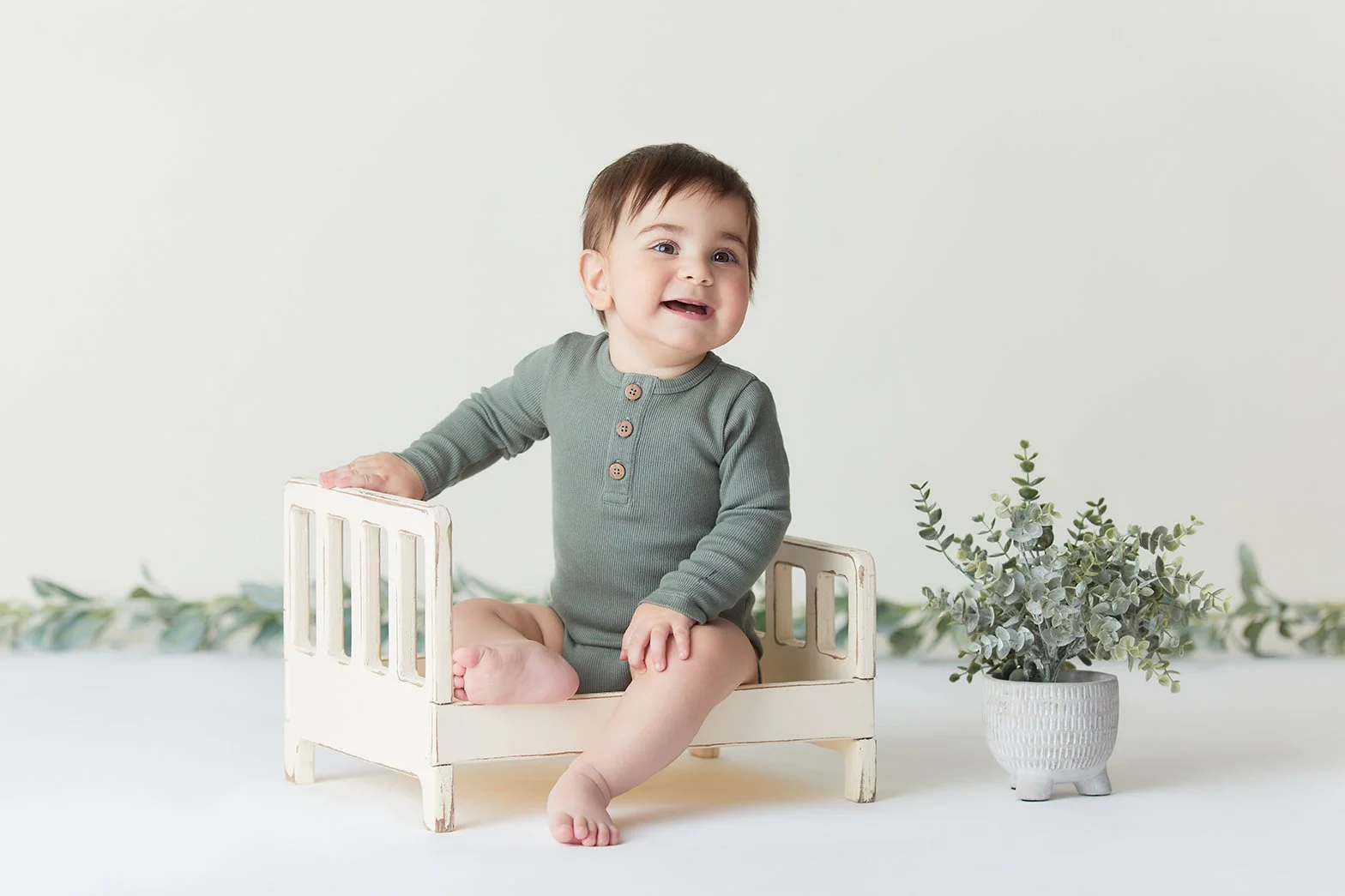 A young baby with brown hair, wearing a green long-sleeve onesie with buttons, sitting on a small white wooden bed with one leg hanging off. There is a white pot with green foliage to the right of the baby, and a leafy green garland in the background