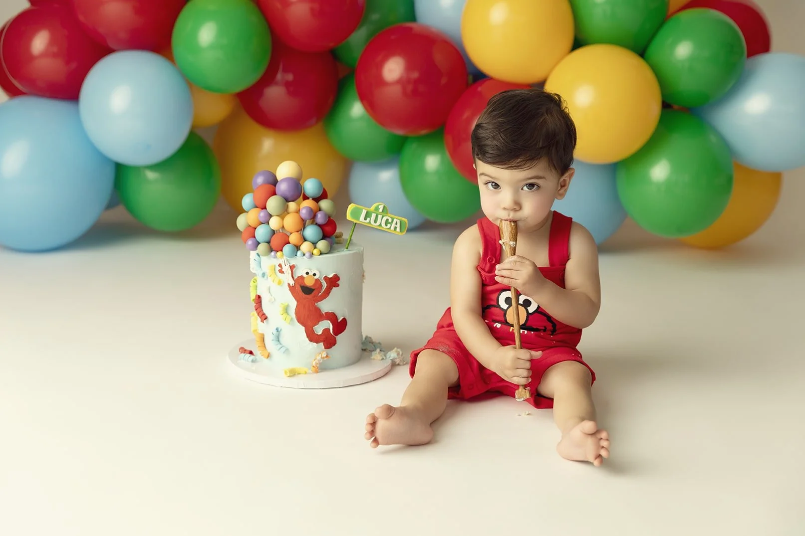 Young boy sitting on the floor in front of colorful balloons, holding a cupcake, with a cartoon character and a decorated birthday cake nearby.