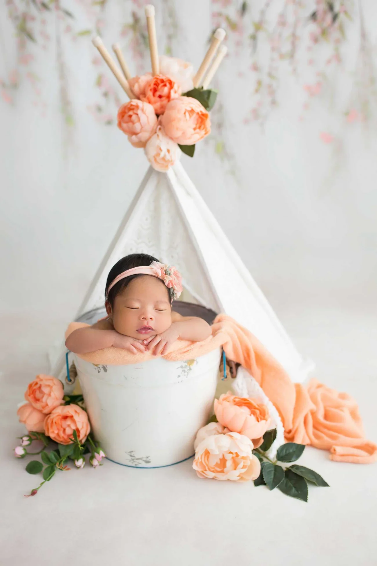A sleeping baby girl with a pink headband and flower accessory rests her chin on her folded arms over a soft peach blanket inside a white bucket. The setup includes a small white teepee decorated with pink flowers and greenery, with additional peach 