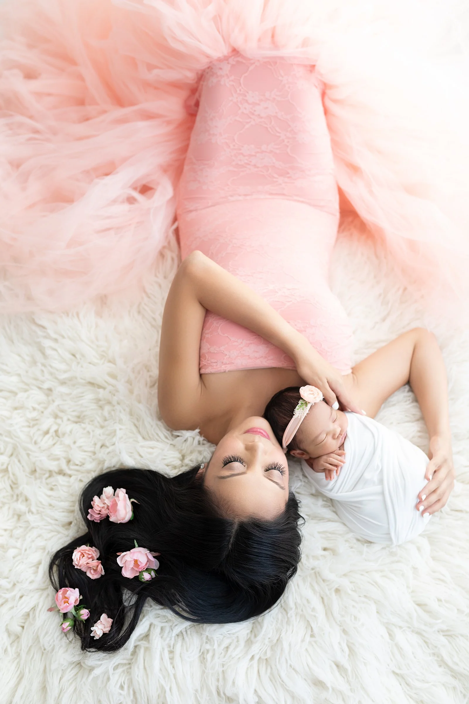 A woman and a baby lying on a fluffy white rug. The woman is wearing a pink lace dress and has long black hair adorned with pink flowers. The baby is wrapped in a white blanket and has a pink headband with a flower, sleeping peacefully.