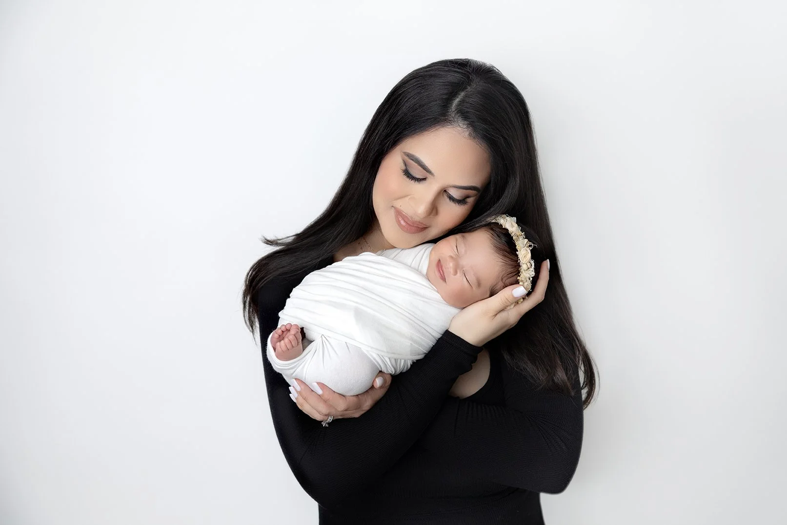 A woman with long dark hair holding a sleeping newborn wrapped in a white blanket, against a white background.