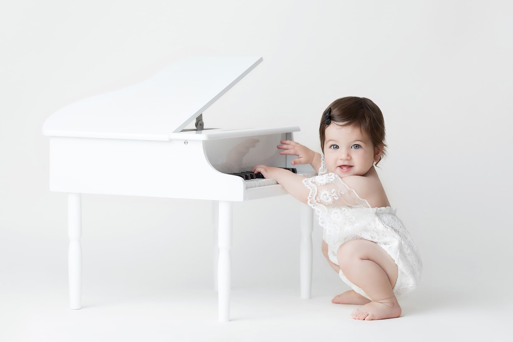 A smiling baby girl with dark hair and blue eyes, wearing a white lace dress with short sleeves, is squatting next to a small white toy piano, touching the keys. The piano has a closed lid and four legs, and the baby is looking at the camera with a j