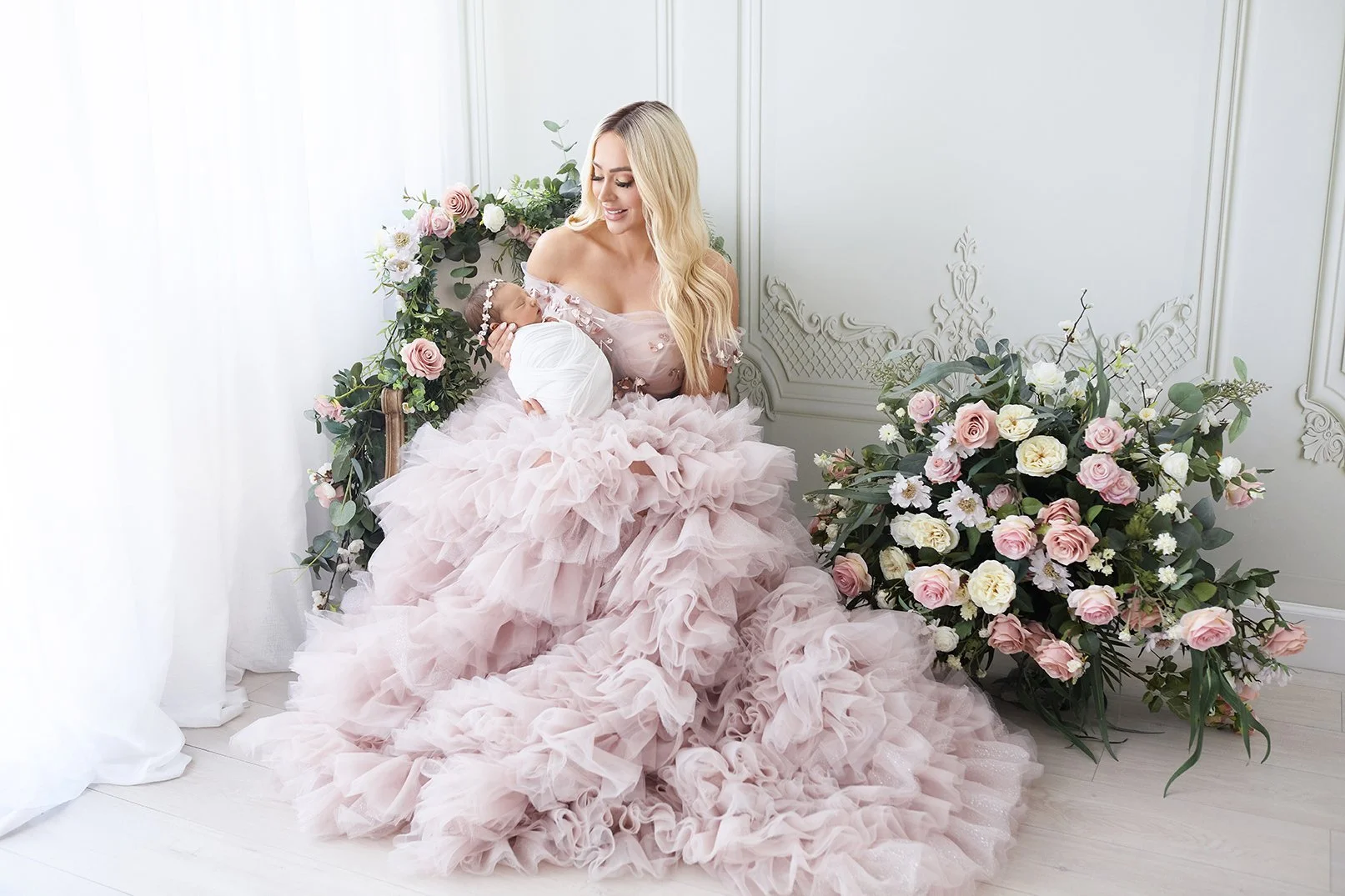 A woman in a pink ruffled dress holding a newborn baby wrapped in white blanket, sitting near floral arrangements in a bright room.