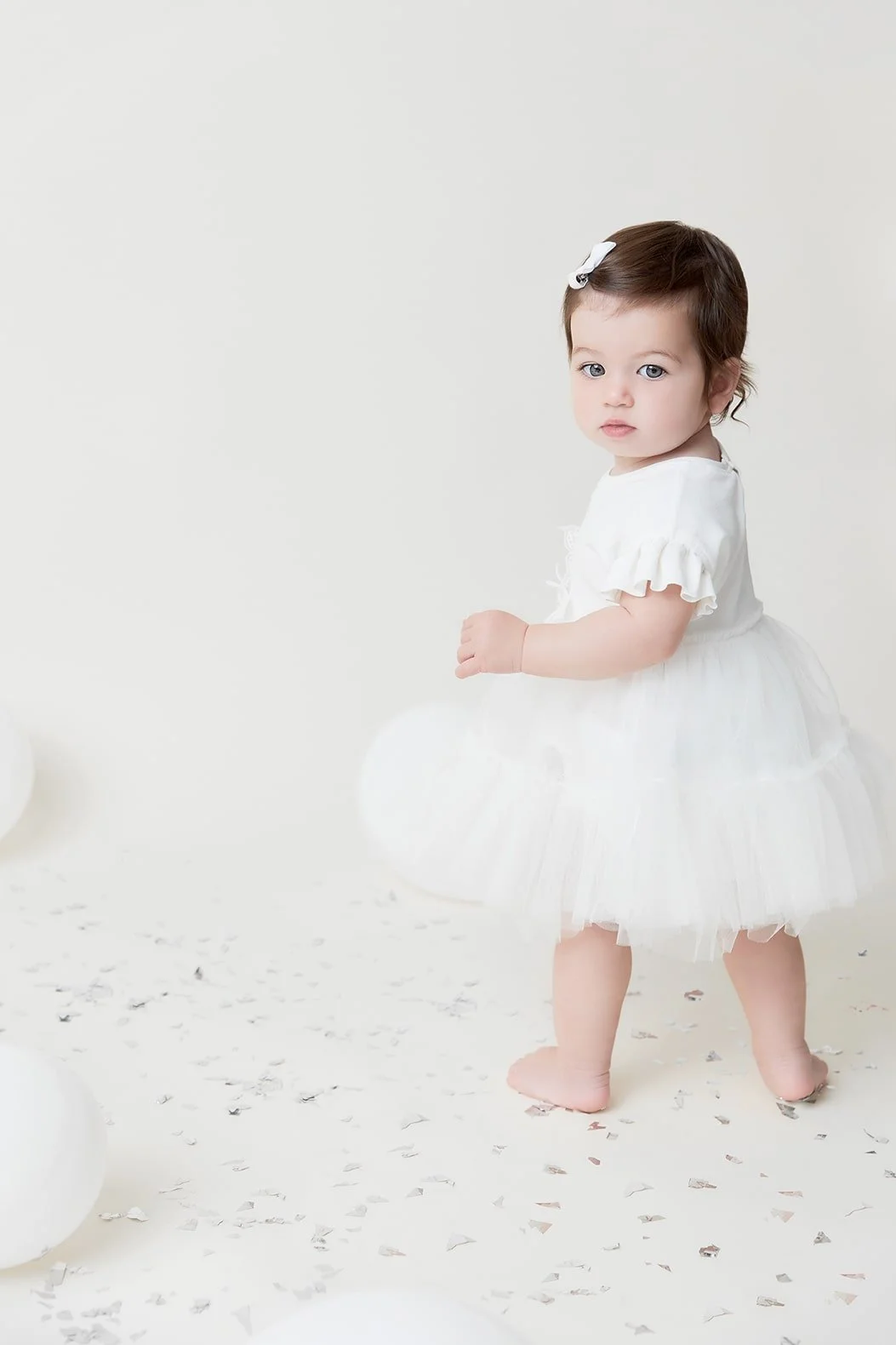 A young toddler girl with brown hair and blue eyes, wearing a white dress with ruffled sleeves and a bow in her hair, standing barefoot on a white floor with scattered silver confetti.