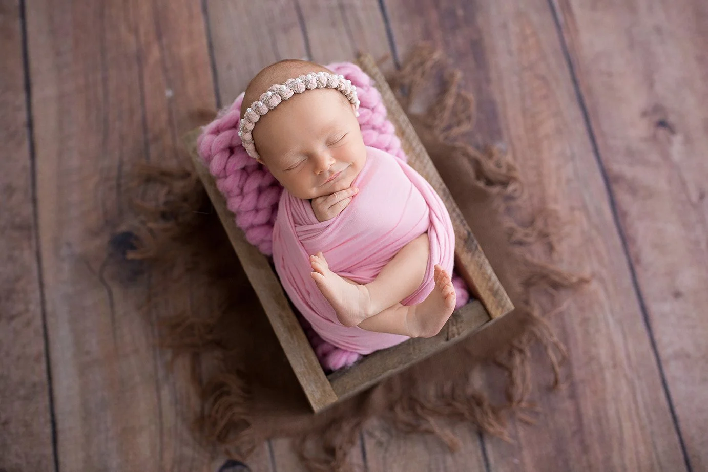 A sleeping newborn baby wrapped in pink cloth, resting in a small wooden box with a pink knitted blanket and a beige beaded headband.