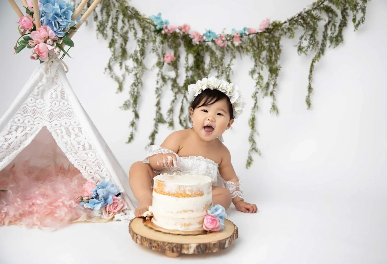 A happy baby girl wearing a white lace dress and a flower crown, sitting in front of a small, semi-naked birthday cake with pink and blue flowers, next to a decorative tent with flowers and lace, against a white background with hanging greenery and p