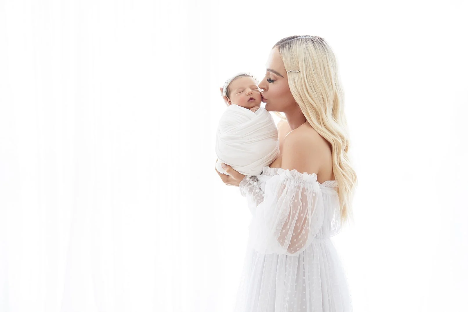 A woman with long blonde hair wearing a white off-shoulder dress, holding a newborn wrapped in white swaddle, while kissing the baby's nose against a white background.