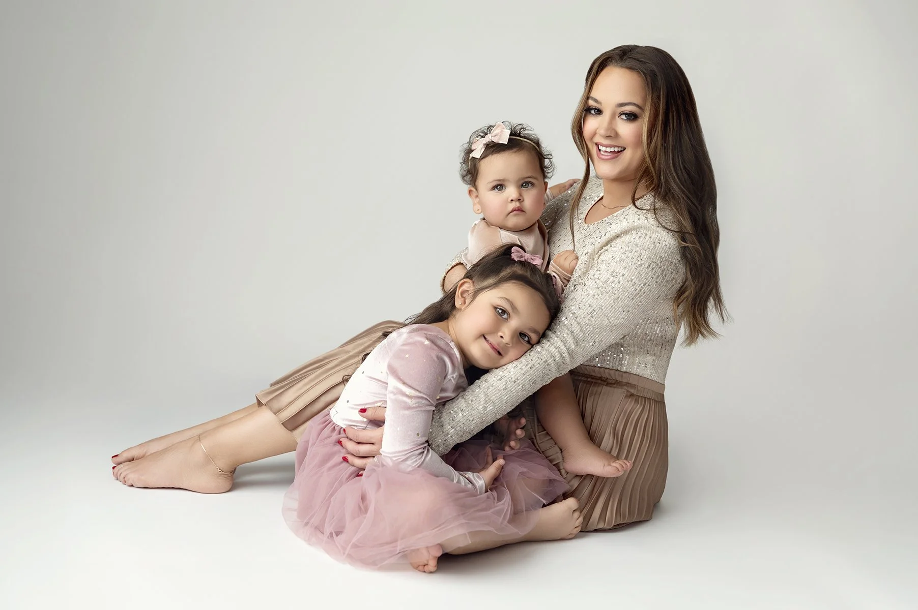A woman with long brown hair smiling sitting with three young girls against a plain white background.