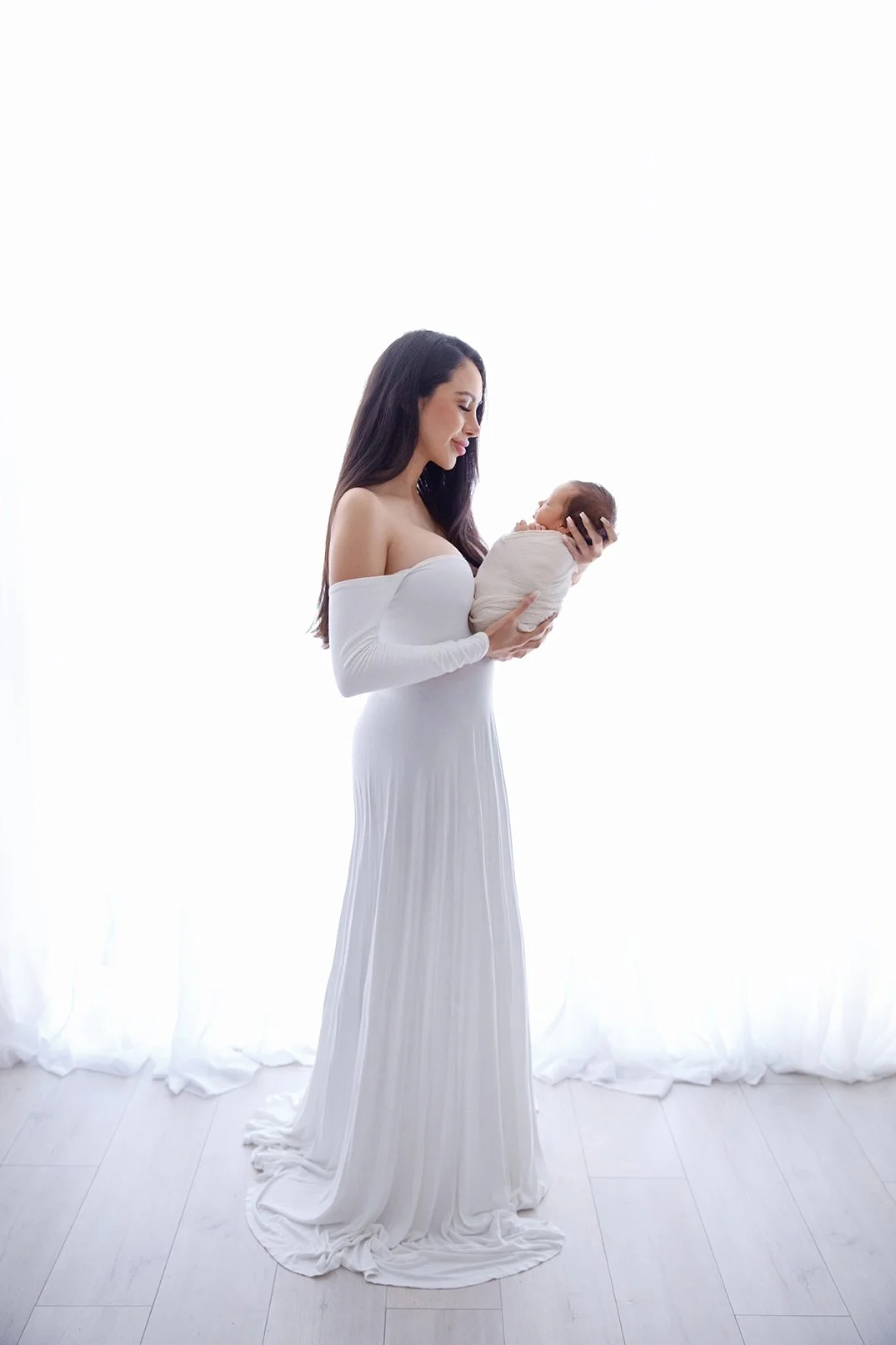 A woman in a long white dress holds a newborn wrapped in white cloth, standing in front of a bright white background.