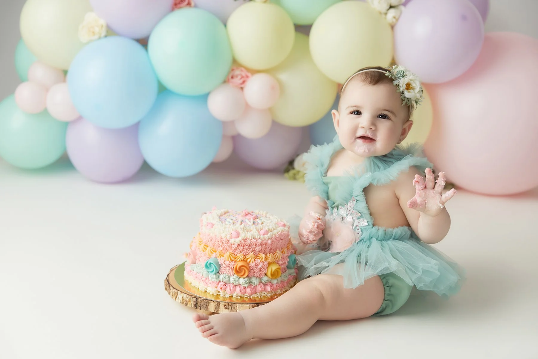 A baby girl celebrating her first birthday with a colorful cake and pastel balloons in the background.