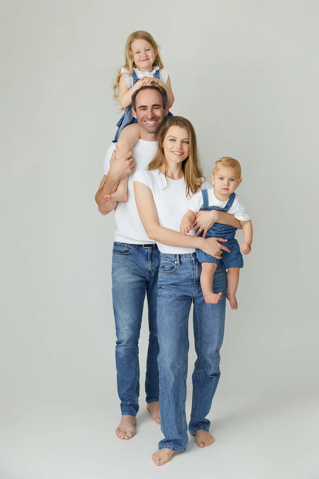 A happy family of four, with a man, woman, and two young children, posing together barefoot against a plain light background. The girl is sitting on the man's shoulders, and the boy is held by the woman.