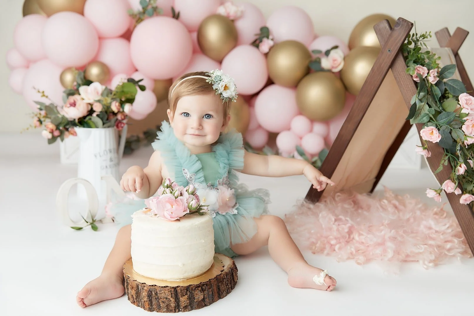 A young girl with blue eyes and a floral headband celebrating her first birthday, sitting on the floor next to a small white cake with pink flowers, in a decorated setting with pink and gold balloons, flowers, and themed decorations.