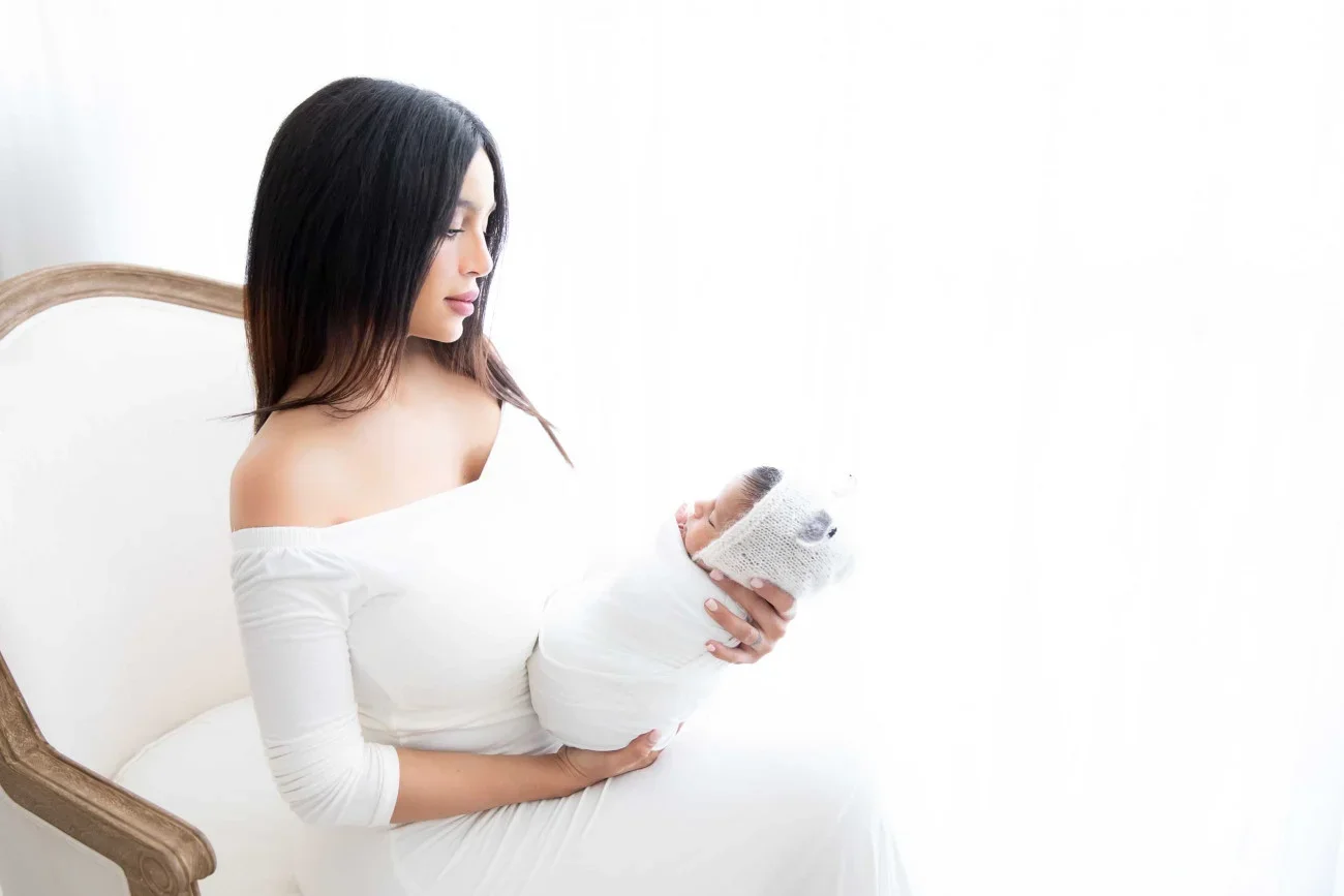 A woman with long dark hair holding a newborn baby wrapped in a white blanket, sitting on a white chair in a bright room.