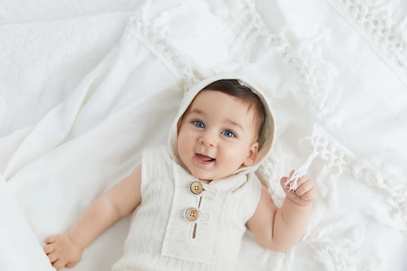 Baby lying on white blankets wearing a cream-colored sleeveless hoodie with wooden buttons, smiling and holding a woolen tassel.