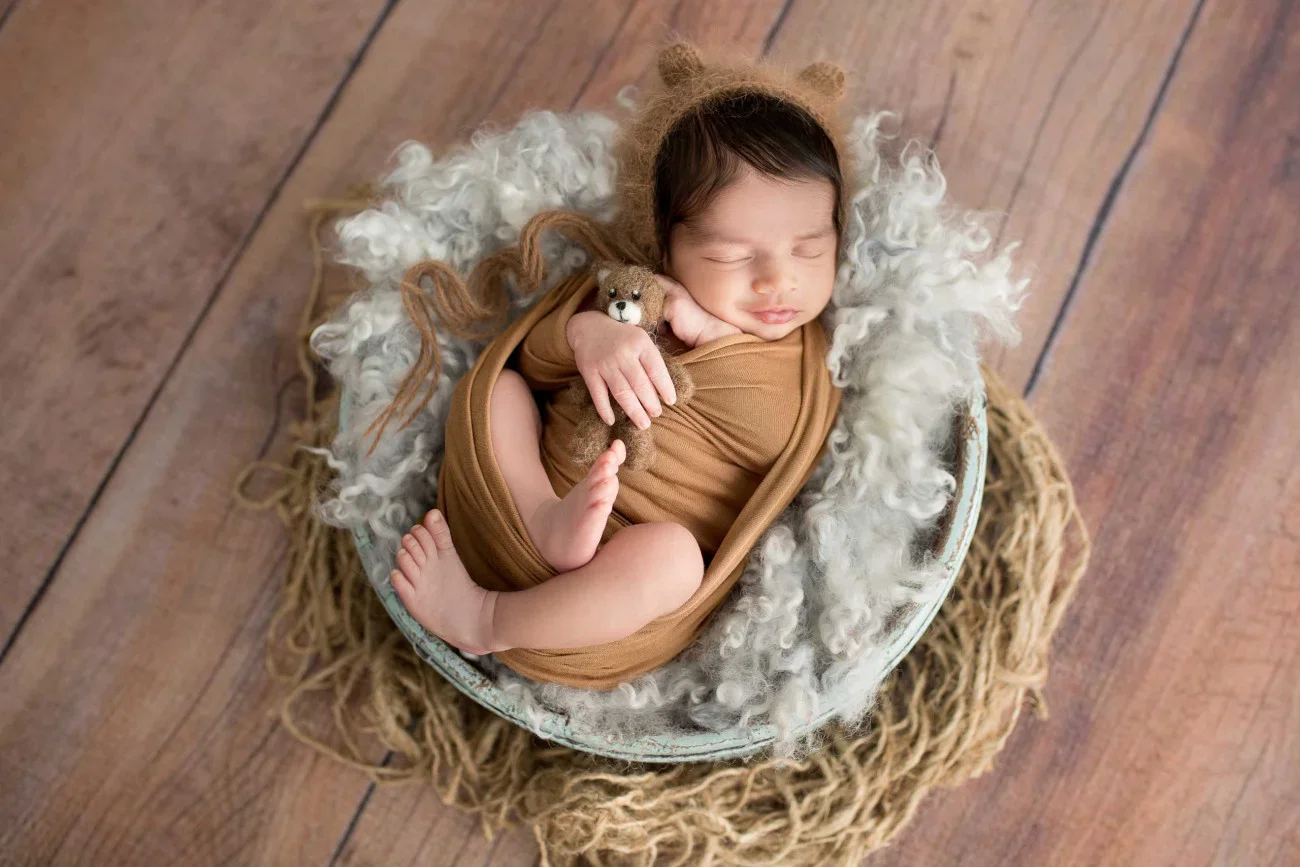 A sleeping newborn baby wrapped in a brown blanket, wearing a bear hat, and holding a small teddy bear, lying in a round basket surrounded by white fluffy material on a wooden floor.