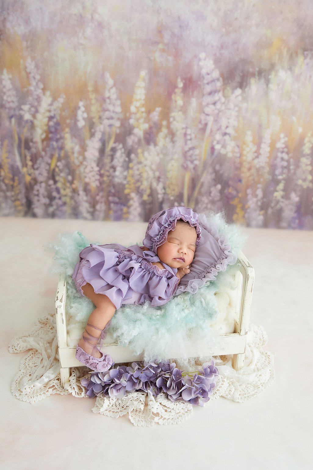 A baby dressed in purple with a ruffled bonnet lying on a small white wooden bed, surrounded by pastel-colored flowers and lace, with a lavender flower backdrop.
